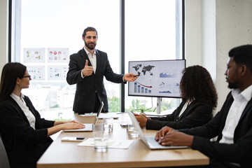 Businessman reports to colleagues standing near graphs and charts on monitor, screen, display. Conference room with professionals discussing financial data. Team meeting emphasizing business strategy.