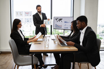 Team engaged in presentation, using laptops, and analyzing financial data. Business team meeting with professionals discussing documents and charts in modern office setting.