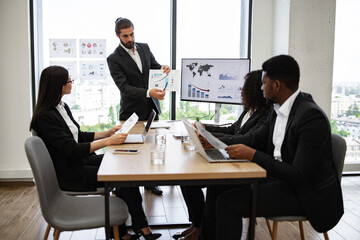 Business team in formal meeting discussing charts and graphs with laptops and documents. Professional presentation in modern office setting with large windows displaying cityscape view.