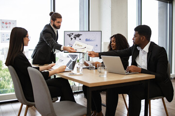 Businessman hands out paper tasks or results of company's activities to colleagues. Professional corporate team collaboratively working on project. Office setting with laptops and presentation screen.