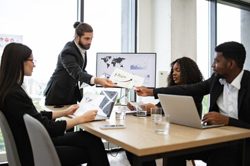 Businessman hands out paper tasks or results of company's activities to colleagues during meeting. Team discussing reports charts and business strategies. Office environment with professional setting