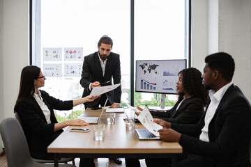 Diverse business team discussing financial graphs in modern office. Colleagues in formal attire collaborating on company strategy. Professional work environment with charts and statistics displayed.