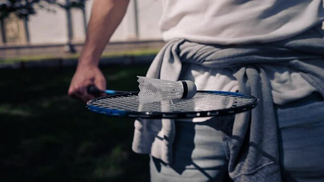 outdoors in a park with trees young handsome brunette guy in jeans and a white T-shirt, a brunette stuffing a shuttlecock warming up before playing bambinton