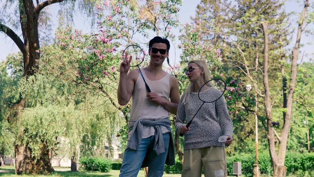 outdoors in a park with trees young brunette guy stands next to a blonde girl bambinton rackets best friends relaxing together