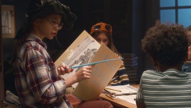 Side shot of girl in wizards hat and with magic stick holding big folder with tabletop game rules and managing board game process while playing with diverse friends at home in evening