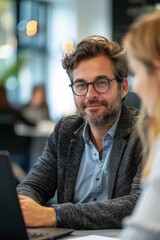 Fototapeta premium A confident businessman with glasses working on a laptop in a contemporary office environment, engaging in a conversation.