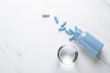 Blue pill capsules spilling out of bottle and glass of water on white background, top view