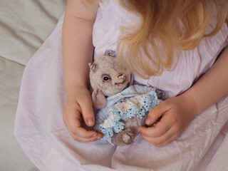 Handcrafted Teddy Lamb in Blonde Toddler Girl’s Hands Against a Pastel Pink Cotton Dress Backdrop. A horizontal close up.