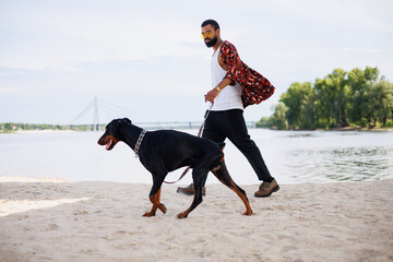 Stylish african american man in sunglasses walking with doberman dog on beach