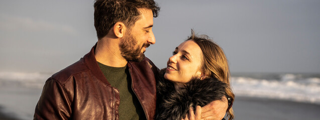 Horizontal banner with couple smiling and embracing at the beach during sunset. They share a moment of love and connection with the waves in the background, creating a warm and romantic scene.