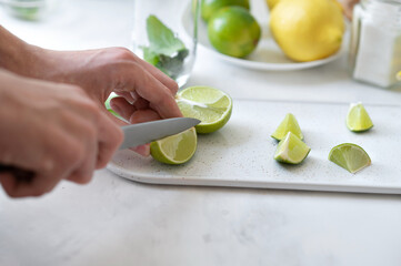 Hands slicing limes on pieces on cutting board, limes, lemon and mint on white table background, soft natural light, fresh juicy citrus fruit, preparing ingredients for mojito recipe