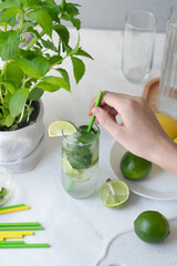 Hand holding drinking straw, putting in glass with mojito cocktail drink, fresh green limes and mint on home kitchen table background, summer homemade refreshing beverage, lifestyle
