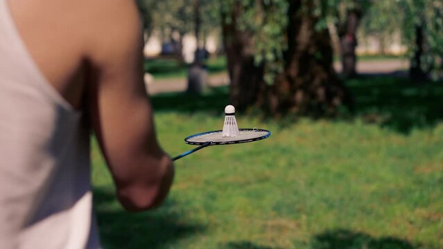 close up outdoors in a park with trees young handsome brunette guy in jeans and a white T-shirt, a brunette stuffing a shuttlecock warming up before playing bambinton