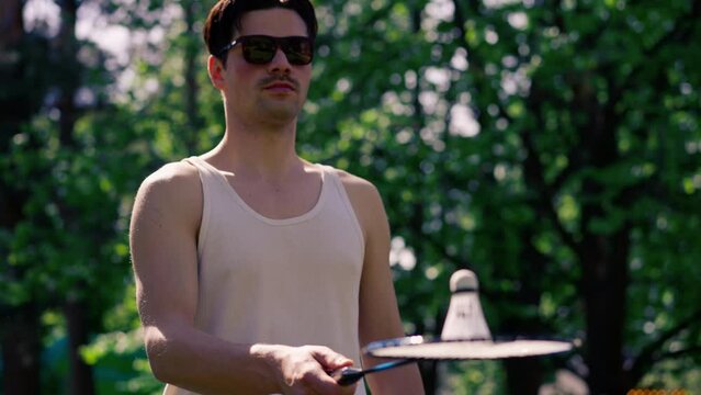outdoors in a park with trees young handsome brunette guy in jeans and a white T-shirt, a brunette stuffing a shuttlecock warming up before playing bambinton