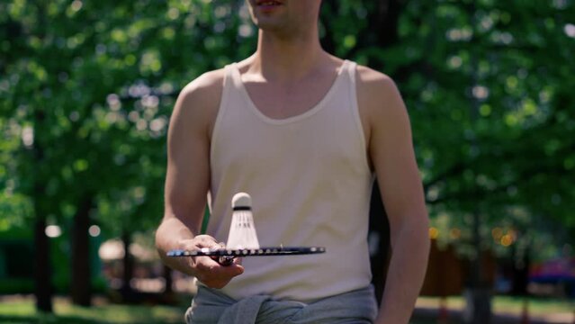 outdoors in a park with trees young handsome brunette guy in jeans and a white T-shirt, a brunette stuffing a shuttlecock warming up before playing bambinton