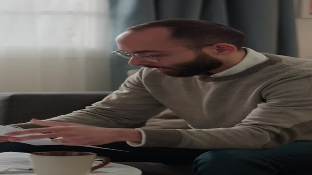 Medium vertical shot of young Middle Eastern man managing domestic expenses, opening envelope, taking out new bill, reading it, clenching fist and swearing with anger
