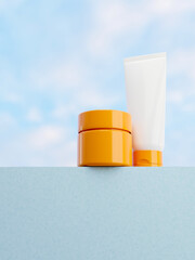 A jar of cream and a white tube mockup on a blue pedestal with a blue sky in the background.