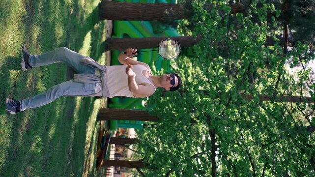 vertical video outdoors in a park with trees young handsome brunette guy in jeans and a white T-shirt, a brunette stuffing a shuttlecock warming up before playing bambinton