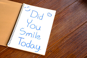 Text of Did You Smile Today? write on the white paper of the notebook an encouraging article or question on a brown wooden background.