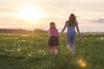 Mother and her little daughter running holding hands across the beautiful green grassland at sunset.
