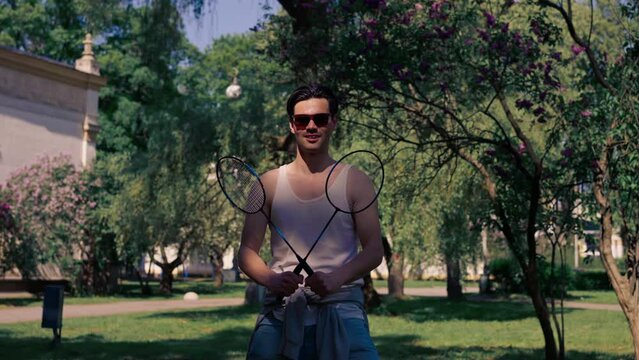on the street in the park among the trees young brunette guy in a white T-shirt is fooling around with two rackets for playing bambinton