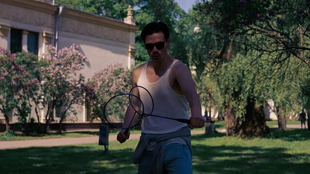 on the street in the park among the trees young brunette guy in a white T-shirt is fooling around with two rackets for playing bambinton