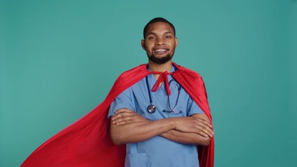 Smiling male nurse portraying superhero wearing red cape, isolated over studio background. Portrait of cheerful heroic medic posing as hero in costume with mantle, camera B
