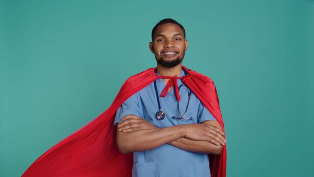 Smiling male nurse portraying superhero wearing red cape, isolated over studio background. Portrait of cheerful heroic medic posing as hero in costume with mantle, camera B