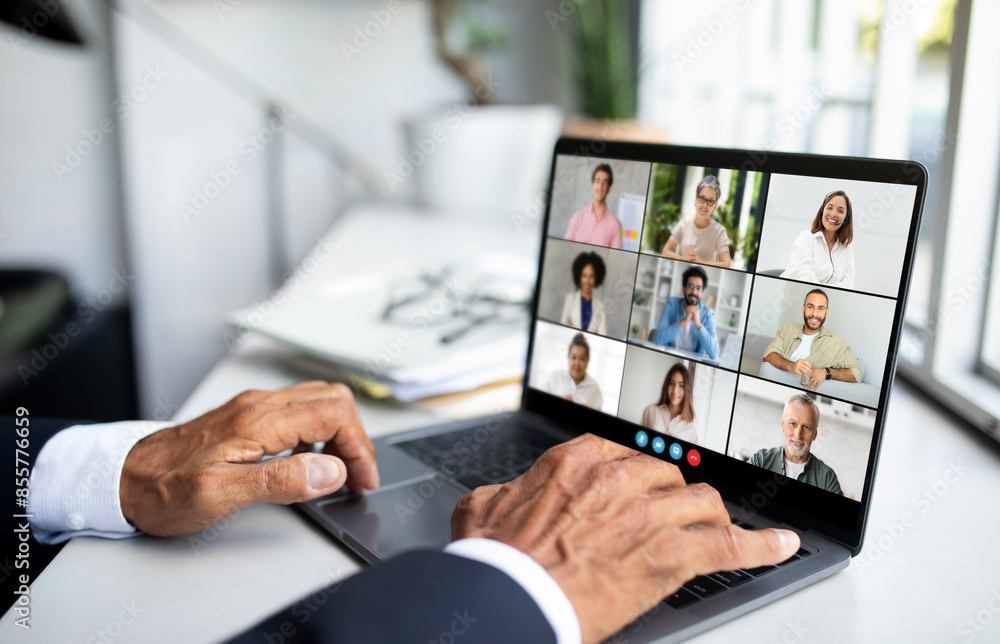 Aufkleber Unrecognizable man is sitting in front of a laptop, engaging in a video call with a group of people displayed on the screen. He appears focused and attentive as he interacts with his virtual audience.