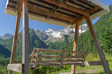 Wooden swing with a canopy to protect from the sun and rain in the mountains for relaxation. Vacation in the mountains in summer. Swing on the background of snow-capped mountain peaks in summer.