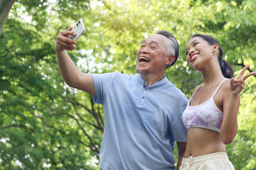Happy elderly Asian man and beautiful daughter taking a selfie together in green summer garden. Grandpa enjoys spending time outdoors with teenage grandchild in park. Senior male using mobile phone.