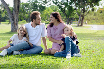 Naklejka premium Happy family sitting on green grass at the park, smiling father, mother, son and daughter spending time together outdoors in beautiful garden on weekend holiday. Parents and kids have fun outside.
