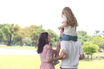 Father carrying cute daughter while taking a walk with mother in beautiful summer garden. Joyful parents piggybacking child girl at green park. Dad and mom play with kid together. Happy family outside