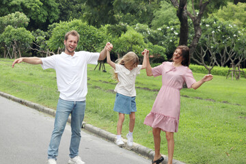 Father, mother and cute kid daughter taking a walk together in beautiful summer garden. Joyful parents hold hands, carry and lift up girl child on the park walkway at green park. Happy family outside.