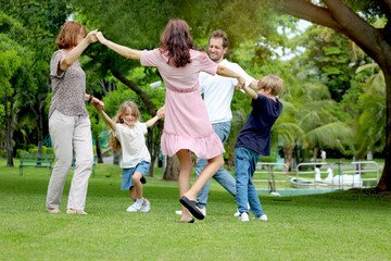 Fototapeta premium Cheerful family hold hands and dances round on green grassy lawn together. Father, mother, daughter girl, son boy and elderly grandma play in outside garden. Happy joyful kid, parents, senior at park.