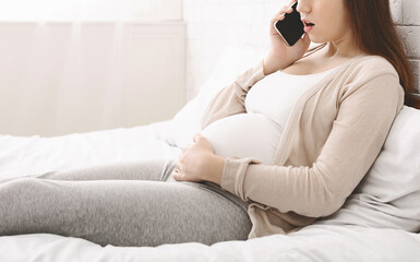 Cropped of pregnant woman is sitting on a bed, engaged in a phone conversation. She appears relaxed and focused as she communicates over the cell phone.