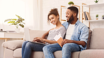 A cheerful African American couple is seen seated closely on a comfortable gray sofa, with the...