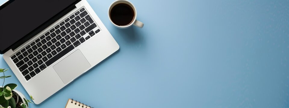 An office desk with a laptop, notebooks, and coffee cup on a blue color background. A panoramic banner background with space for a copy.