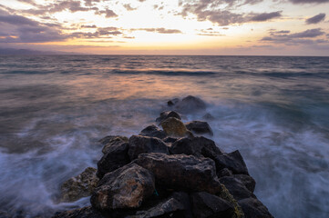 Dramatic sunset over the Mediterranean Sea with rocks in the foreground. Cloudy sky and waves on the beach. 1