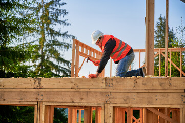 Carpenter constructing two-story wooden frame house near forest. Bearded man hammering nails into...