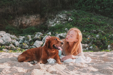 Young red haired woman with freckles lying next to her golden retriever dog on the beach and...