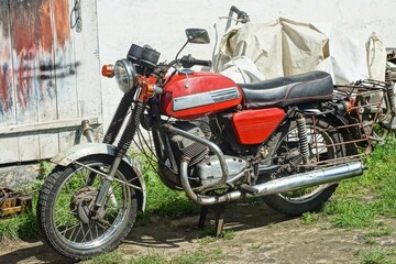 one red retro old rusty heavy with two wheels dirty motorcycle stands on the ground during the day on the street near a white wall and wooden doors