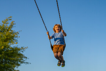 Little boy swinging on chain swing on city kids playground. Swing ride. Cute child having fun on a swing on summer sky background. Blonde little boy swings at kid playground. Child swinging high.