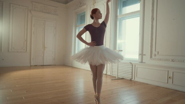 Slender graceful ballerina in tutu dancing in rehearsal room, slow motion. Attractive female ballet dancer exercising in dance hall, standing on parquet with pointe shoes, classical dance performance
