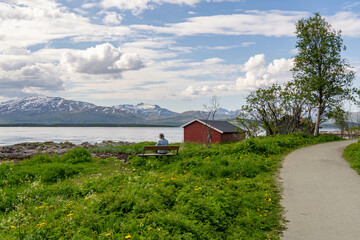 A lonely elderly woman is sitting on the bench and admiring the summer seascape with a traditional Norwegian boathouse, dandelions, mountain, sea, beautiful clouds	