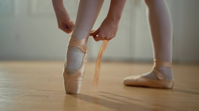 Ballerina tying silk tapes of pointe on slender legs, closeup in ballet hall. Graceful female ballet dancer preparing to rehearsal or performance on stage, details, beauty and art, tender and strong