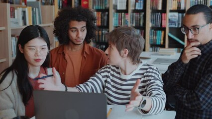 Group of young multi-ethnic students standing around girl explaining them project on laptop monitor in college library