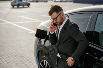 Leaning on the vehicle while it charges, with smartphone. Businessman in suit is near his black car outdoors