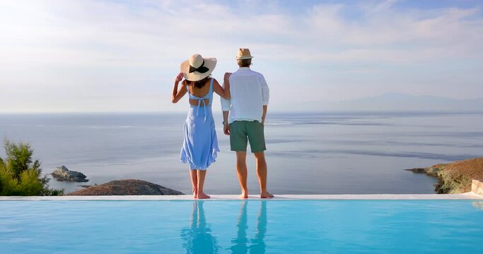 A romantic summer holiday couple stands at the edge of an infinity swimming pool and enjoys the view over the ocean