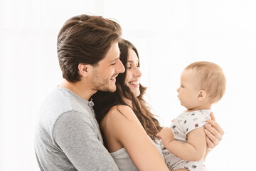 A happy family of three is pictured against a white background. The mother is holding the baby boy, who is looking at his parents. They are all smiling at the baby, showing love and affection.